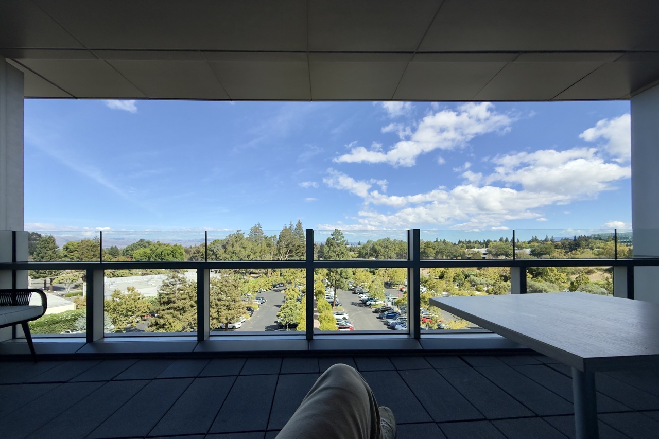 First-person perspective from a shaded balcony with legs propped up on a tiled terrace, looking out past a glass railing onto a sunlit, tree-lined parking lot beneath a bright blue sky filled with puffy white clouds. A white table sits to the right and a dark patio chair is on the left.