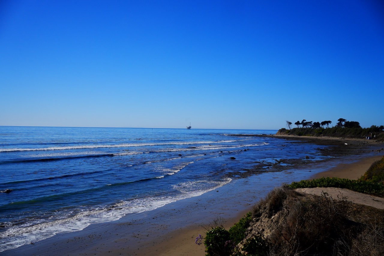 A calm coastline at low tide, an offshore oil platform on the horizon and palm trees lining a distant bluff.