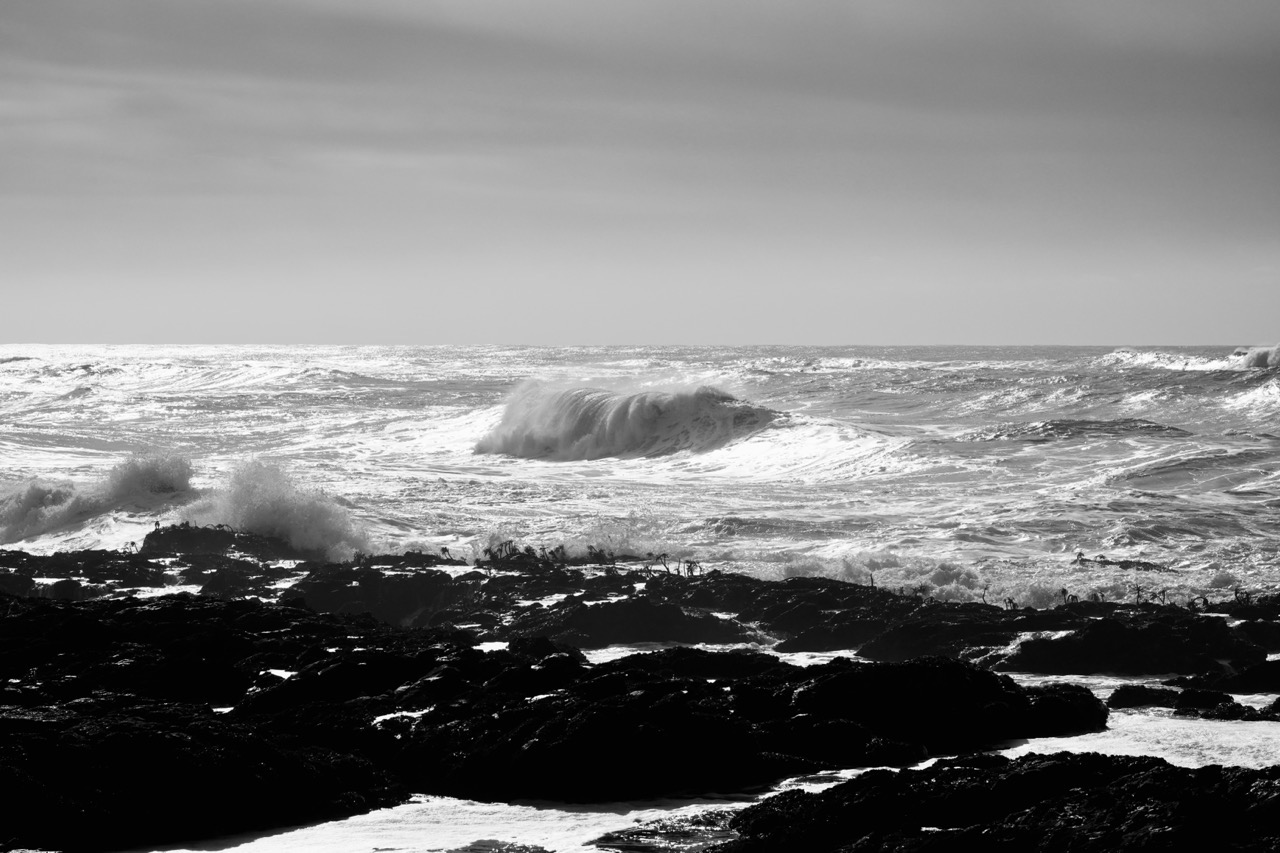 A black-and-white shot of a large wave breaking over dark rocks in rough surf.