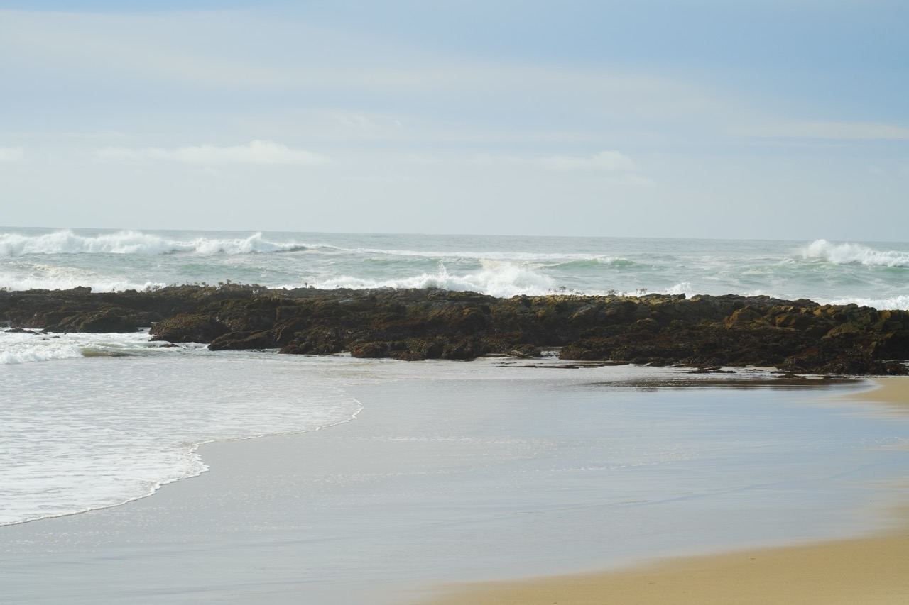 Waves breaking over a dark rocky reef just offshore, with a calm sandy beach in the foreground.