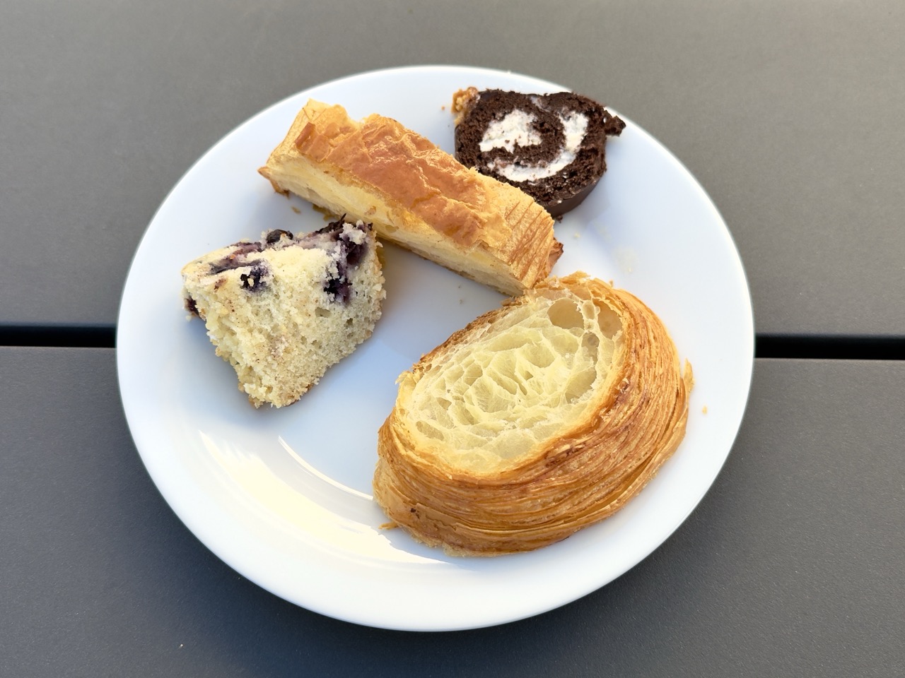 A white plate with cut portions of four pastries: a slice of croissant showing its laminated interior, a piece of blueberry muffin, a slice of pain au chocolat, and an Oh-Oh chocolate swiss roll round.