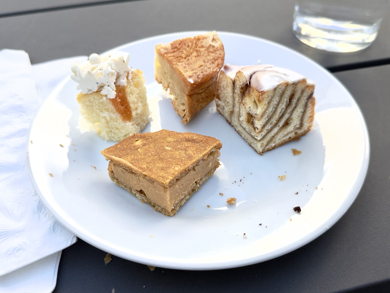 A white plate holding four pastry slices: a peach chiffon cupcake topped with cream and fruit filling, a cross-section of a glazed cinnamon roll, a wedge of Kouign-Amann, and a square of Better Nutter.