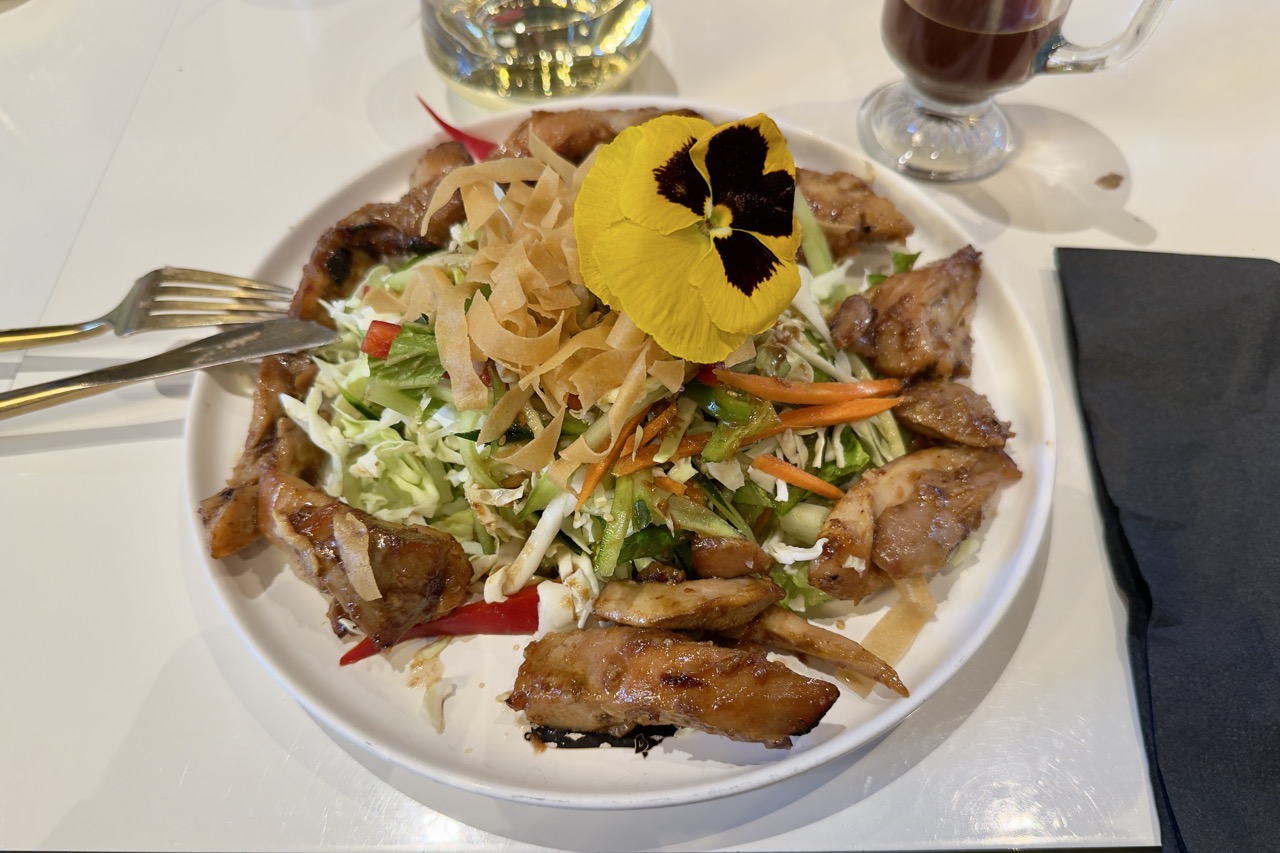 A small fork resting on a glossy white plate of shredded cabbage salad, topped with a yellow pansy and ringed with glazed chicken.