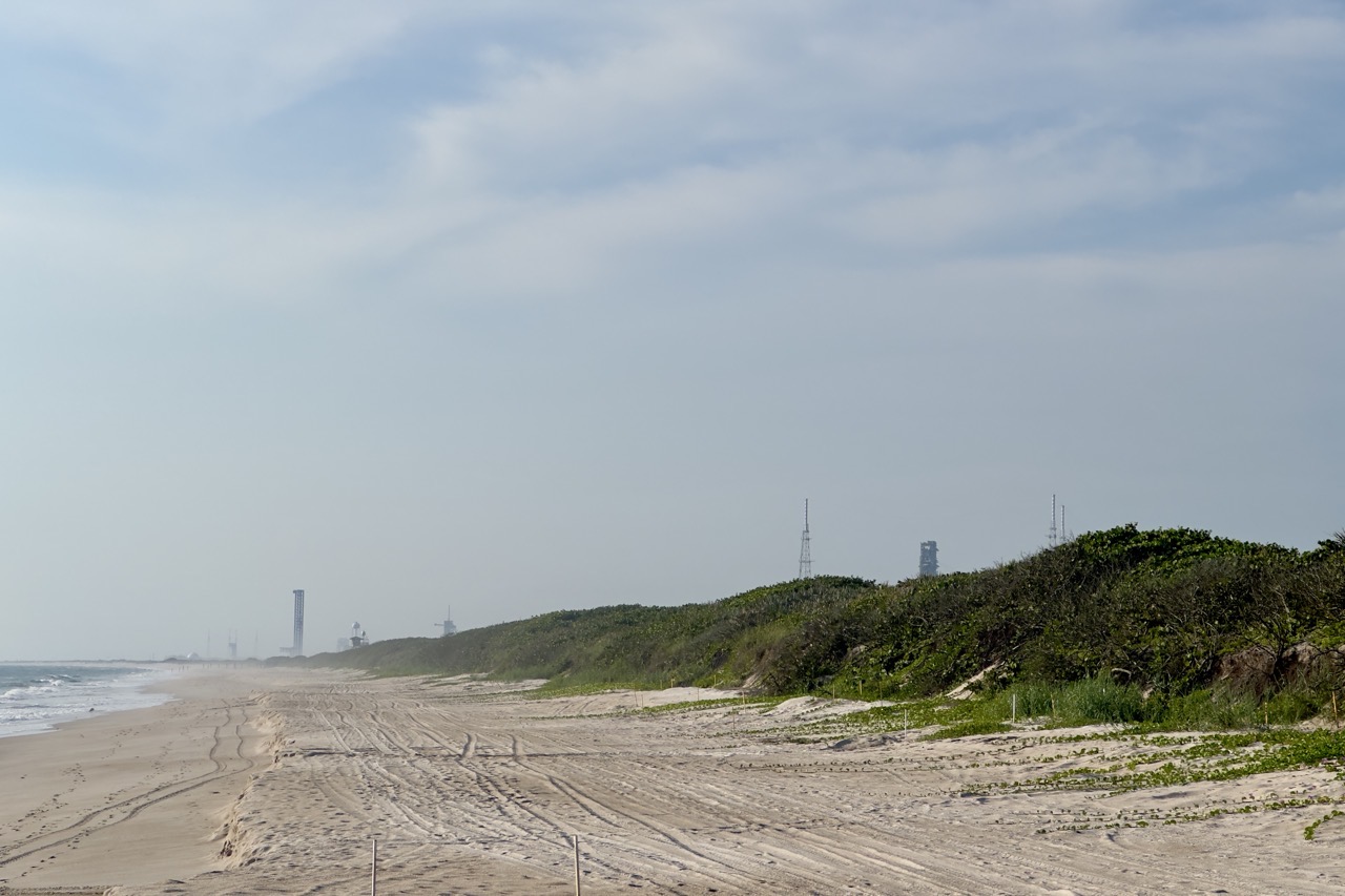 A wide beach with rocket launch towers visible in the distance beyond dune scrub and tire tracks in the sand.
