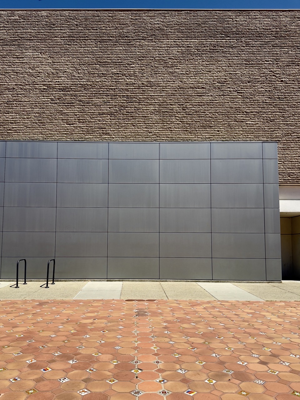 A modern building facade featuring a grid of smooth gray metal panels set against a textured, horizontally ribbed brown masonry wall above. In the foreground, an expansive plaza is paved with hexagonal terracotta tiles accented by small decorative ceramic squares, with two black metal bike racks standing on the concrete walkway at the base of the wall.