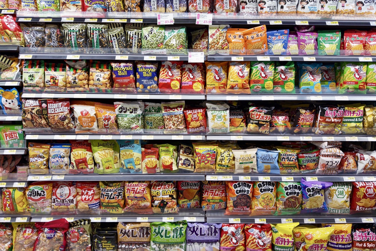 Supermarket shelves lined with bagged chips and rice snacks, Japanese and Korean brands like Calbee, Karamucho, and Takoyaki Ball packed edge to edge.