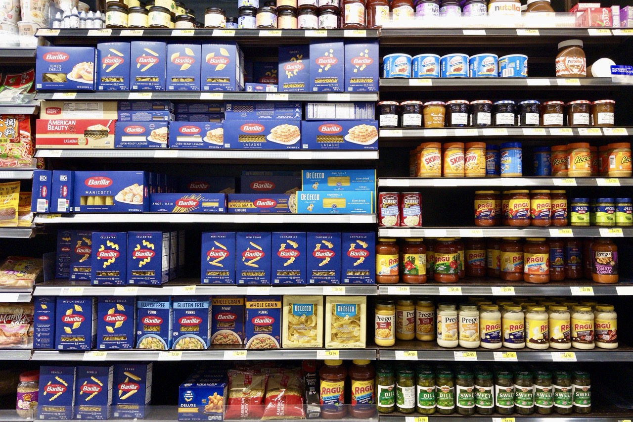 Supermarket shelves stocked with boxed pasta and jarred sauces, Barilla pasta on the left and rows of Ragu, Prego, and peanut butter jars on the right.