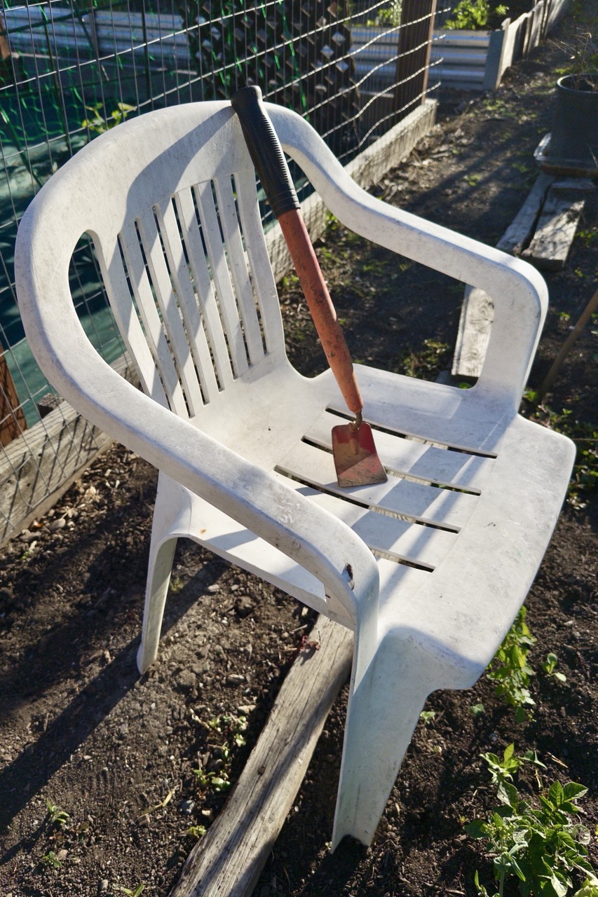 A garden hoe with a thick, hand-shaped wooden handle resting across the seat of a weathered white plastic chair beside a garden bed.