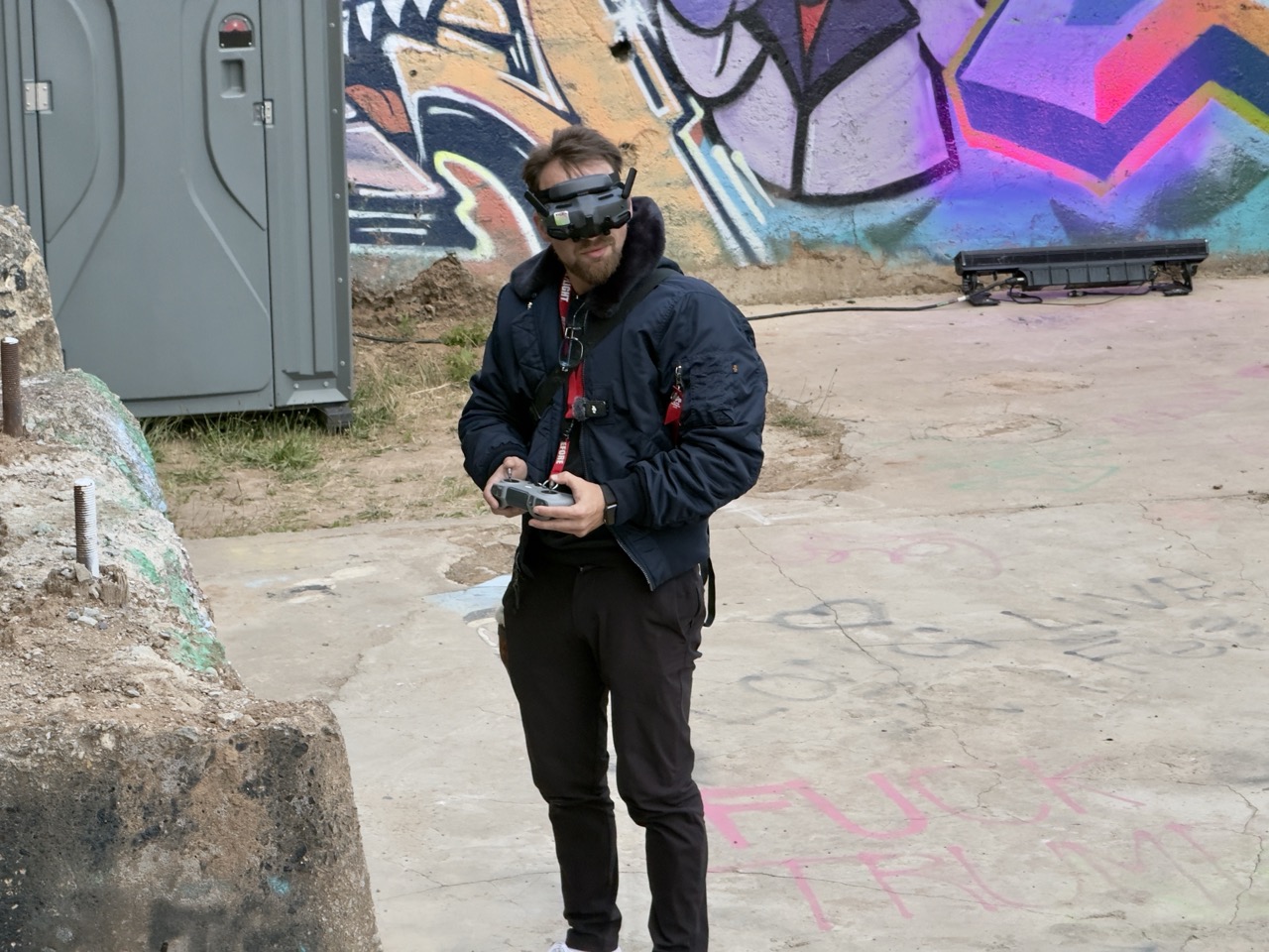 A man wearing FPV video goggles holds a drone controller in front of a graffiti-covered wall.
