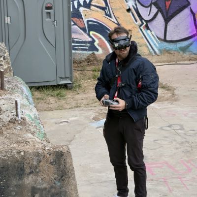 A man wearing FPV video goggles holds a drone controller in front of a graffiti-covered wall.