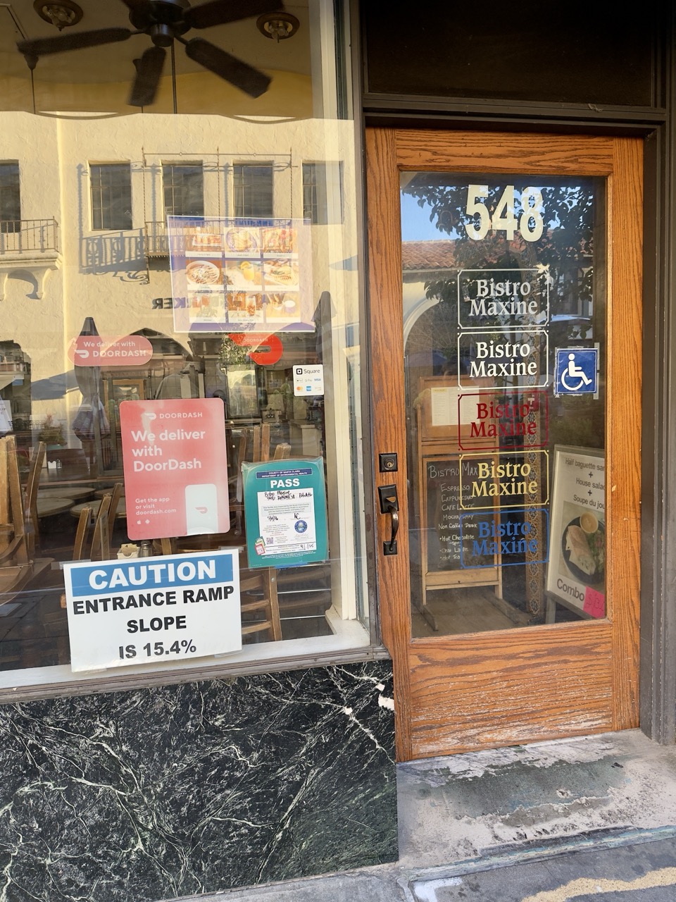 A restaurant entrance for Bistro Maxine, with menus, a DoorDash sign, and a caution sign warning of the ramp's steep slope.