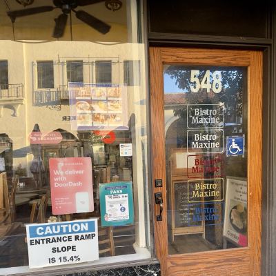 A restaurant entrance for Bistro Maxine, with menus, a DoorDash sign, and a caution sign warning of the ramp's steep slope.