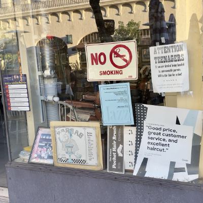 A barbershop window covered in signs, including a no-smoking notice and a tongue-in-cheek 'Attention Teenagers' flyer.