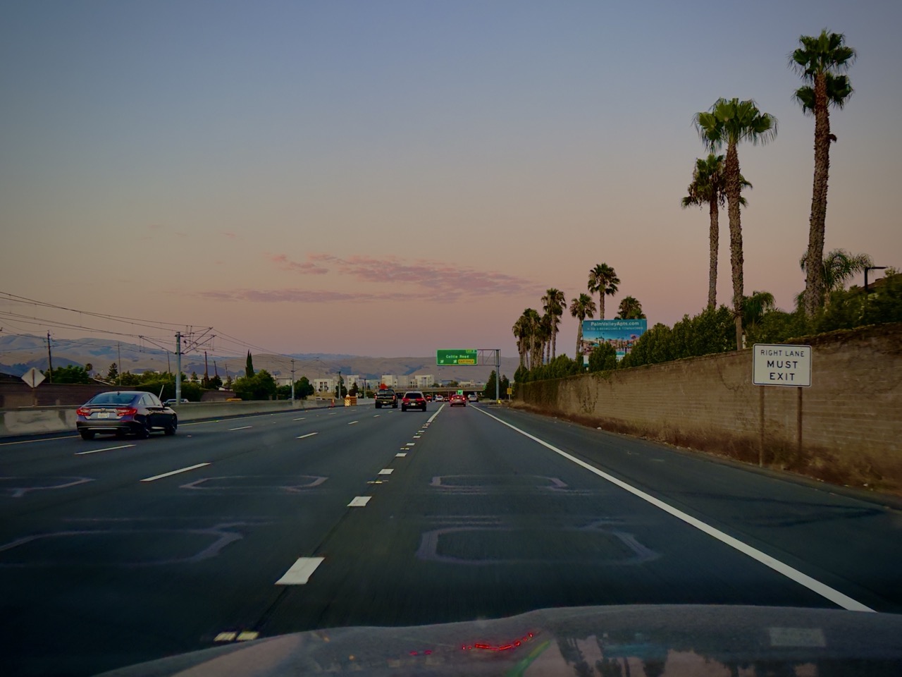 A nearly empty freeway at dusk lined with palm trees, with a 'right lane must exit' sign overhead.