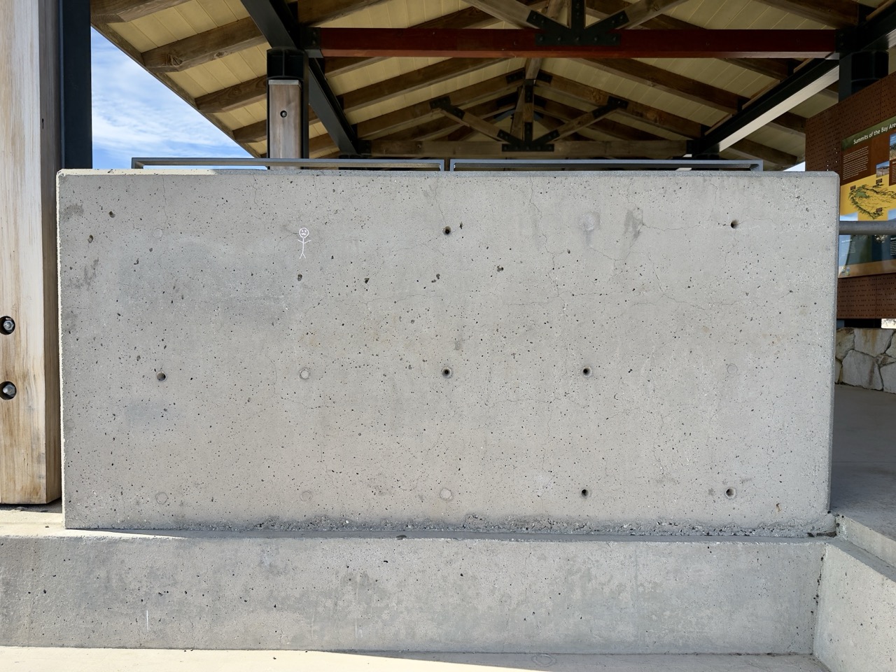 A large concrete pillar beneath a park pavilion's peaked roof, with a tiny stick-figure drawing near its top.