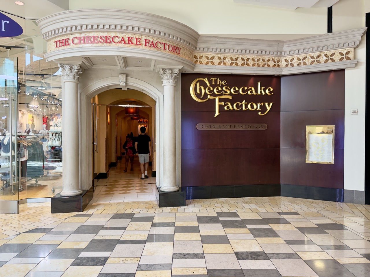 The ornate entrance to a Cheesecake Factory restaurant inside a shopping mall, with shoppers walking past.