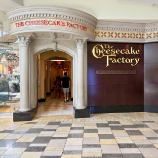The ornate entrance to a Cheesecake Factory restaurant inside a shopping mall, with shoppers walking past.