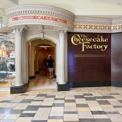 The ornate entrance to a Cheesecake Factory restaurant inside a shopping mall, with shoppers walking past.