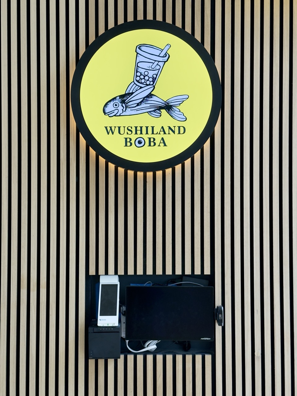 A yellow circular sign for Wushiland Boba featuring a flying fish balancing a boba drink, mounted above a payment terminal.