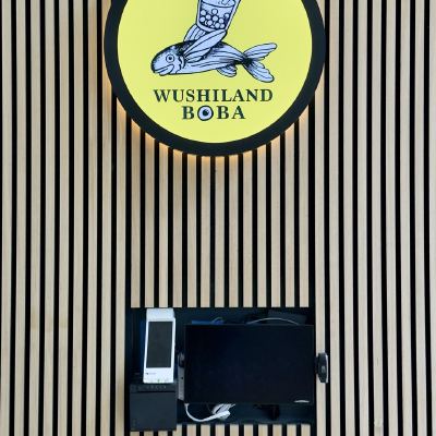 A yellow circular sign for Wushiland Boba featuring a flying fish balancing a boba drink, mounted above a payment terminal.