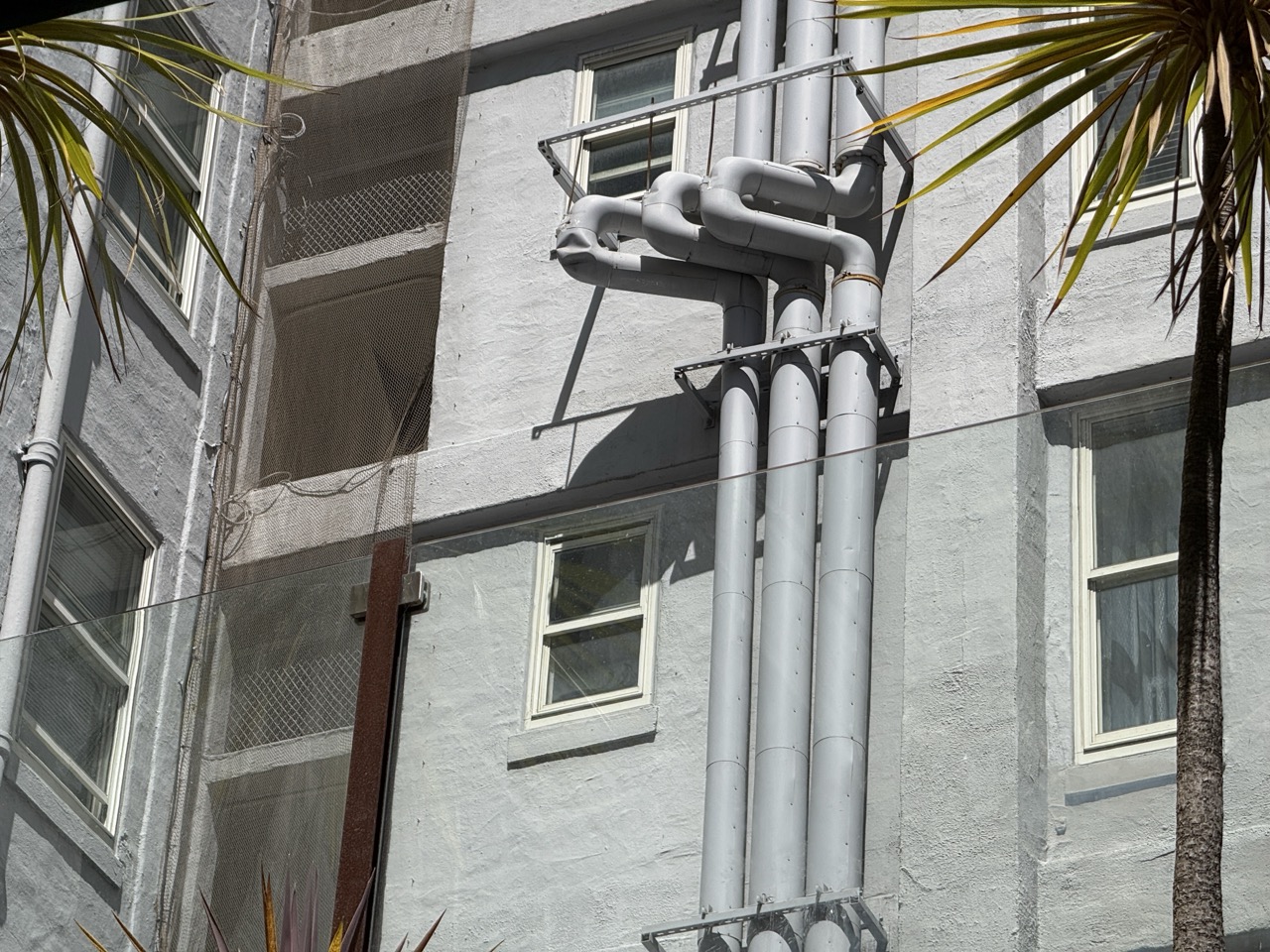 Large gray exhaust pipes running up the side of a stucco building, framed by a palm tree
