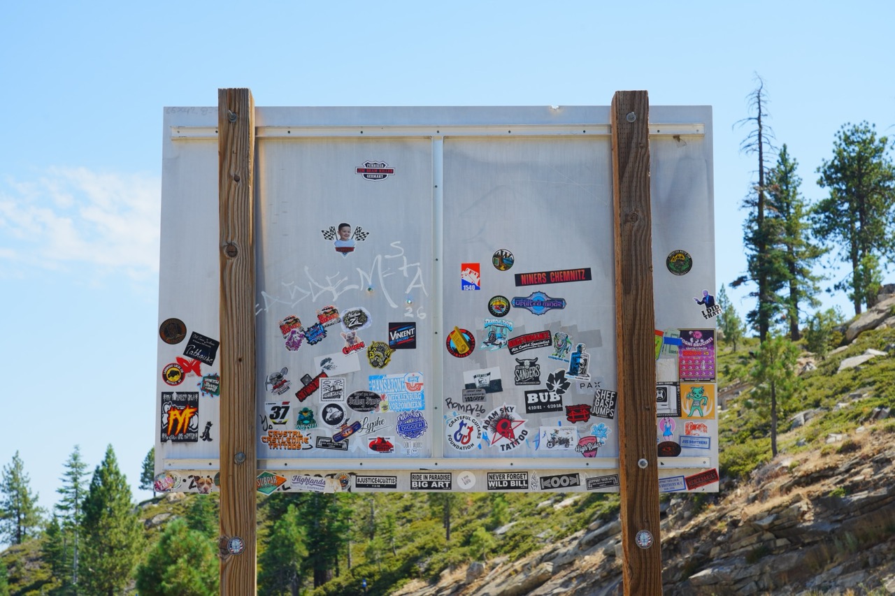 A wooden sign board covered edge to edge with stickers, set against a pine-covered hillside