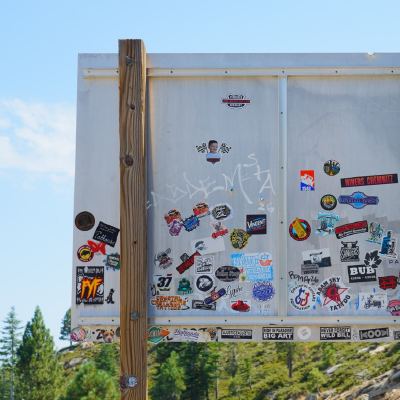 A wooden sign board covered edge to edge with stickers, set against a pine-covered hillside