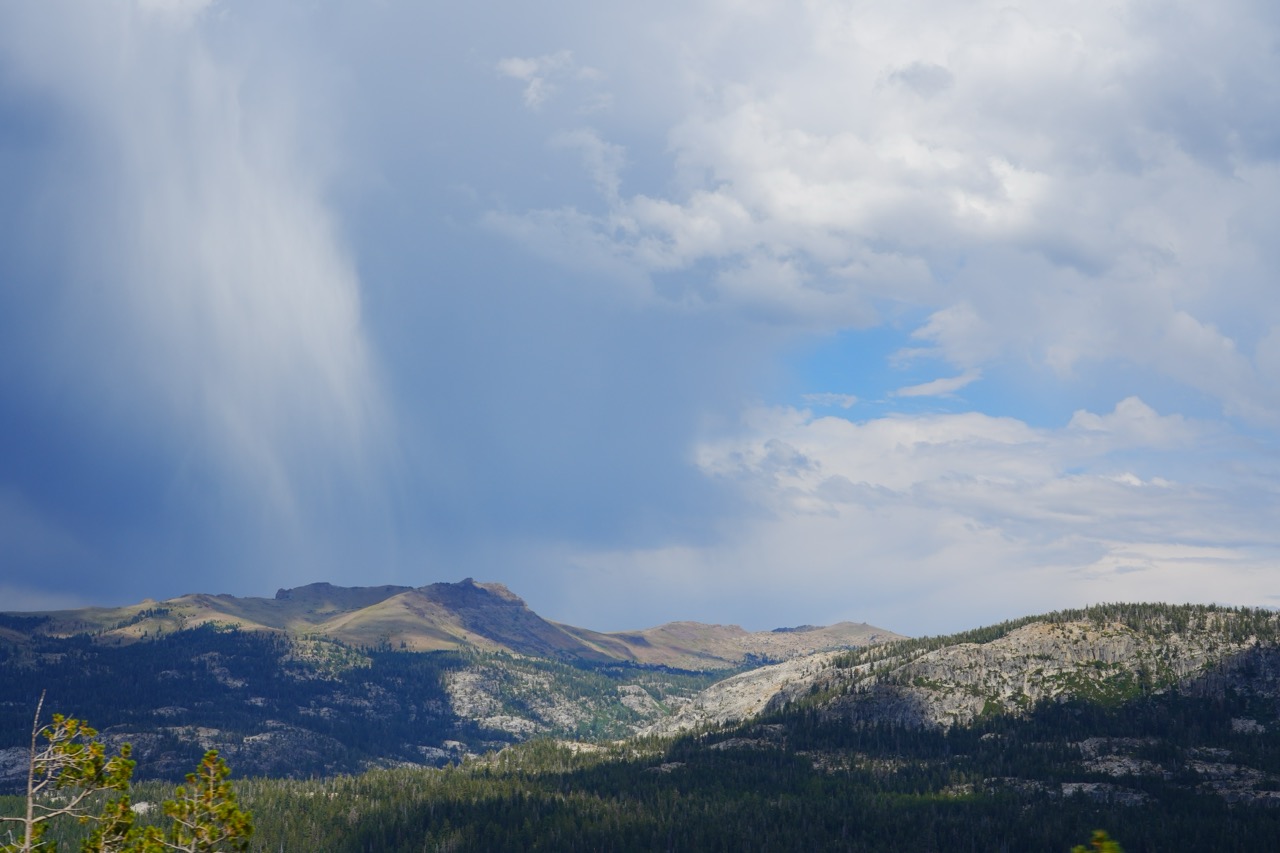 A rain shaft falling from dark clouds over a ridge of granite peaks and forest