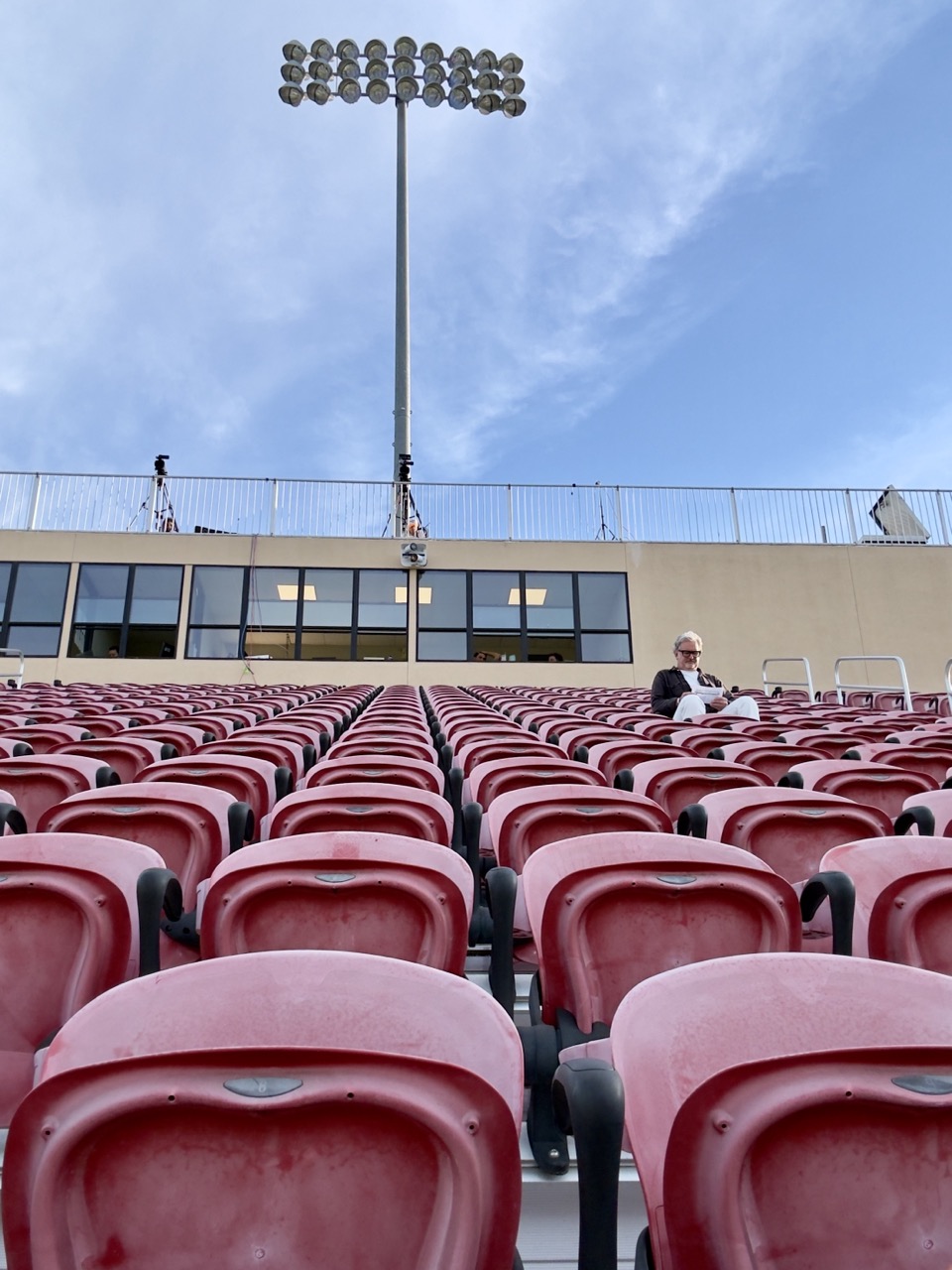 Rows of empty red stadium seats with a single person sitting near the top, under stadium lights