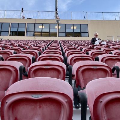 Rows of empty red stadium seats with a single person sitting near the top, under stadium lights
