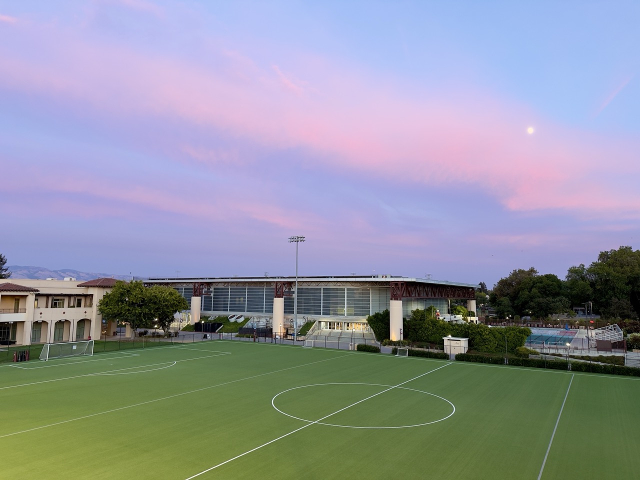 A soccer field at dusk under a pink sky, with the moon visible above a sports building