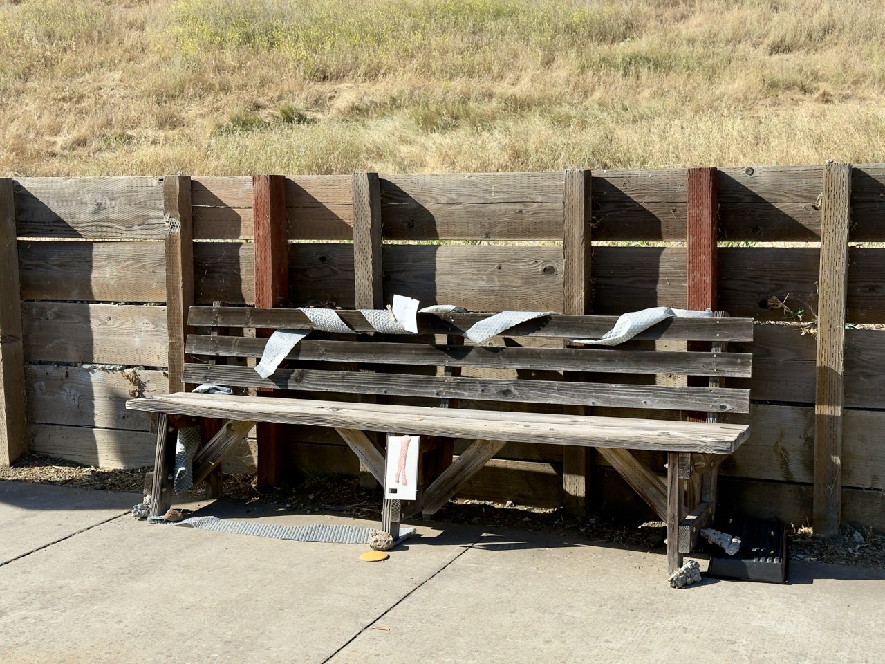A weathered wooden bench against a plank fence, strips of bubble wrap draped over its back, dry grass on the slope behind.