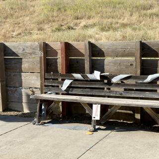 A weathered wooden bench against a plank fence, strips of bubble wrap draped over its back, dry grass on the slope behind.