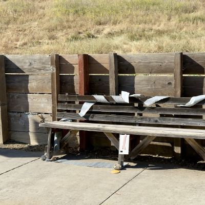 A weathered wooden bench against a plank fence, strips of bubble wrap draped over its back, dry grass on the slope behind.