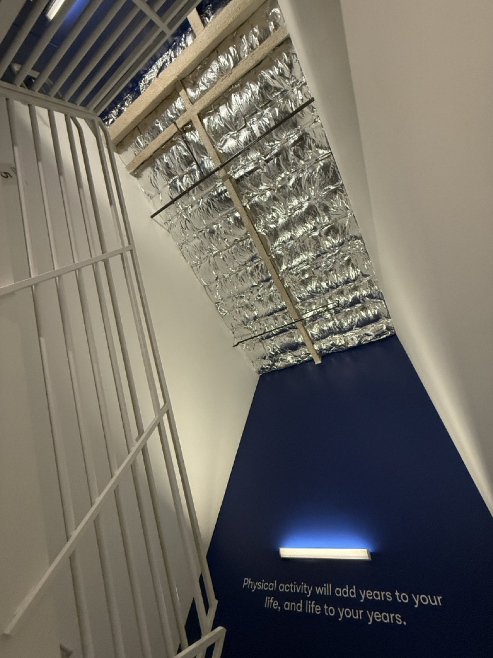 Looking up a narrow stairwell to a ceiling of exposed silver duct insulation, a wall below reading "Physical activity will add years to your life, and life to your years."