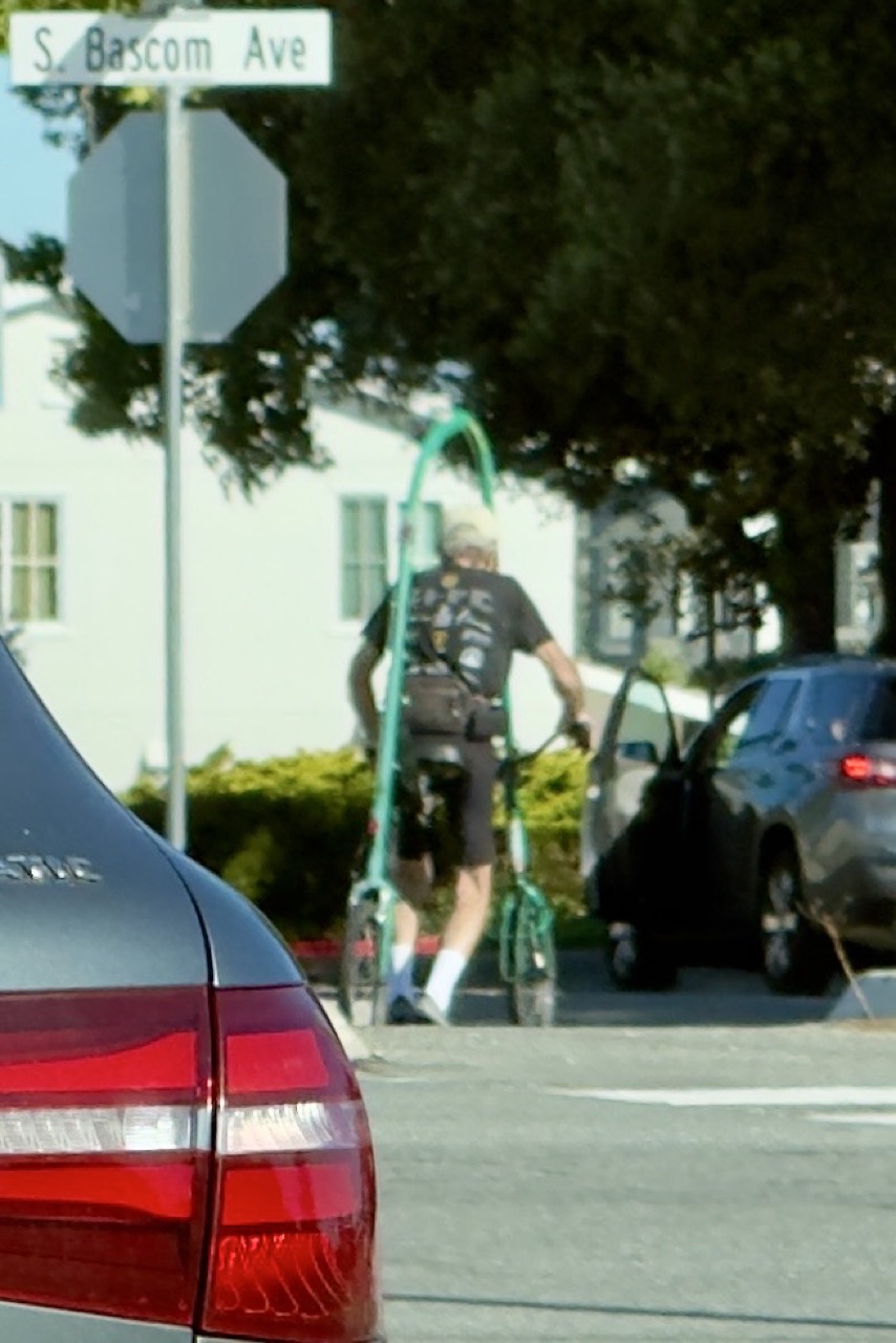 An older man in a cap crossing a street with a green walker frame, an "S. Bascom Ave" sign and parked cars around him.