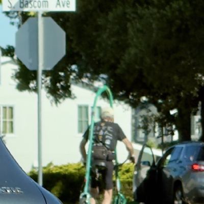 An older man in a cap crossing a street with a green walker frame, an "S. Bascom Ave" sign and parked cars around him.