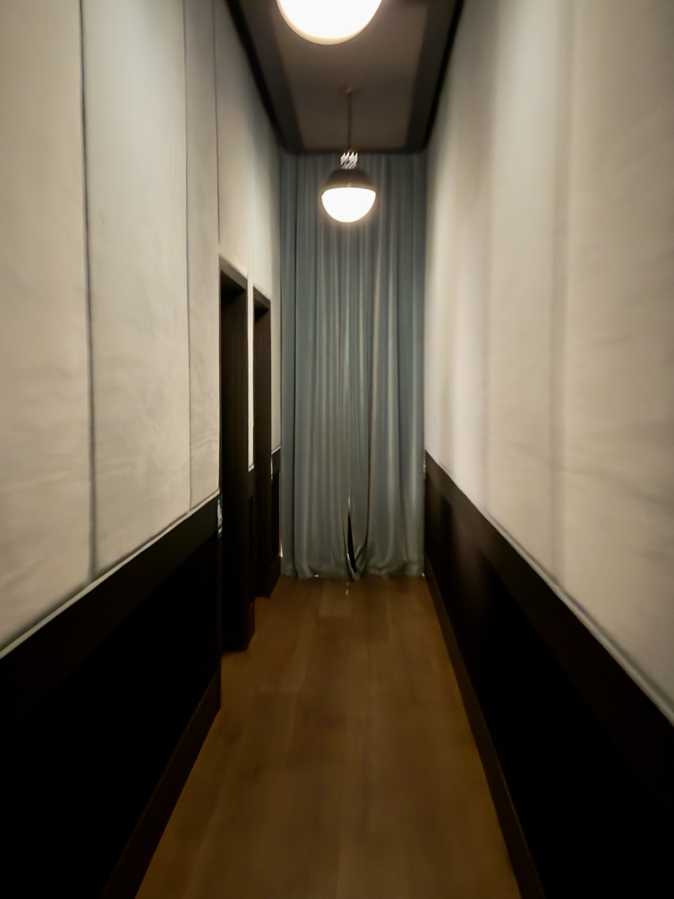 A dim, narrow hallway with pale padded walls and a wood floor, a single globe pendant lighting a grey curtain at the far end.