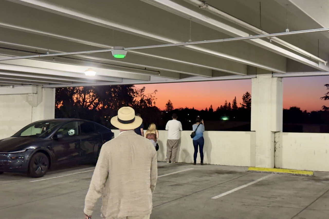 People at the open edge of a parking garage watching an orange sunset over a treeline, a man in a straw hat and linen jacket walking toward them.