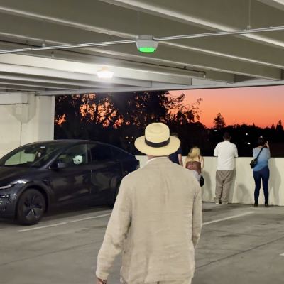 People at the open edge of a parking garage watching an orange sunset over a treeline, a man in a straw hat and linen jacket walking toward them.