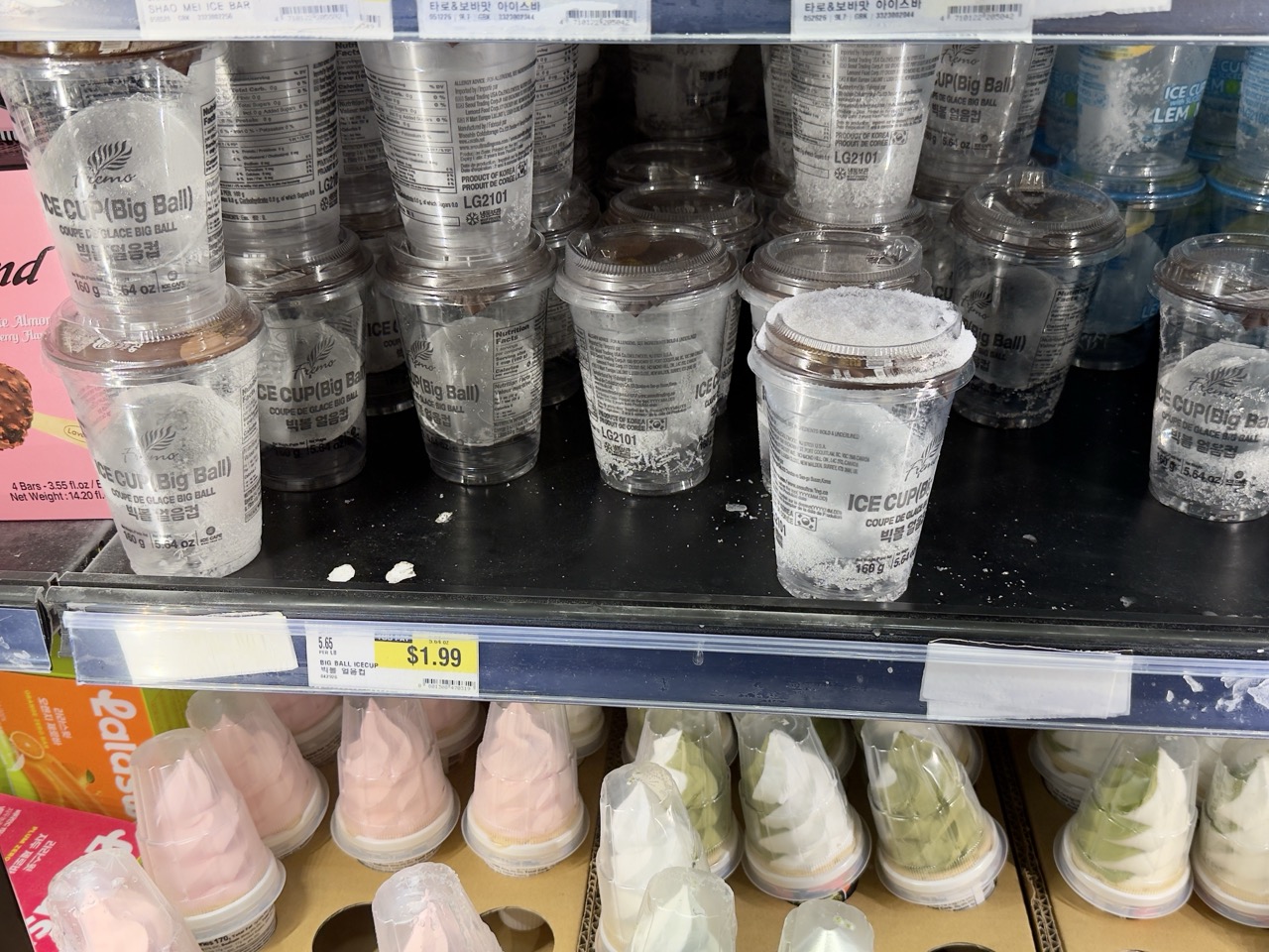 A grocery cooler shelf stacked with clear "Ice Cup (Big Ball)" cups, soft-serve cups on the shelf below and a $1.99 price tag.