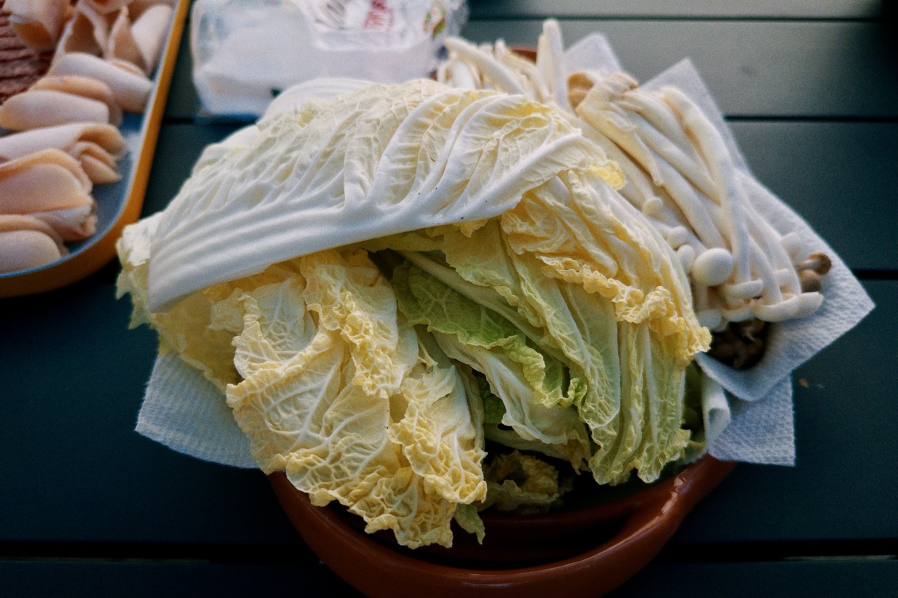 A halved napa cabbage and enoki mushrooms on paper towels in a terracotta bowl, a plate of sliced fish behind, on a slatted table.