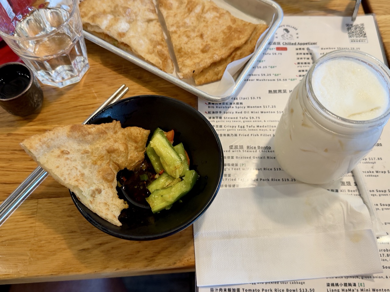 Overhead view of a wooden restaurant table featuring a black bowl with a wedge of flaky scallion pancake and marinated cucumber slices, a metal tray of additional pancake wedges, and a glass jar of soy milk resting on a bilingual menu.