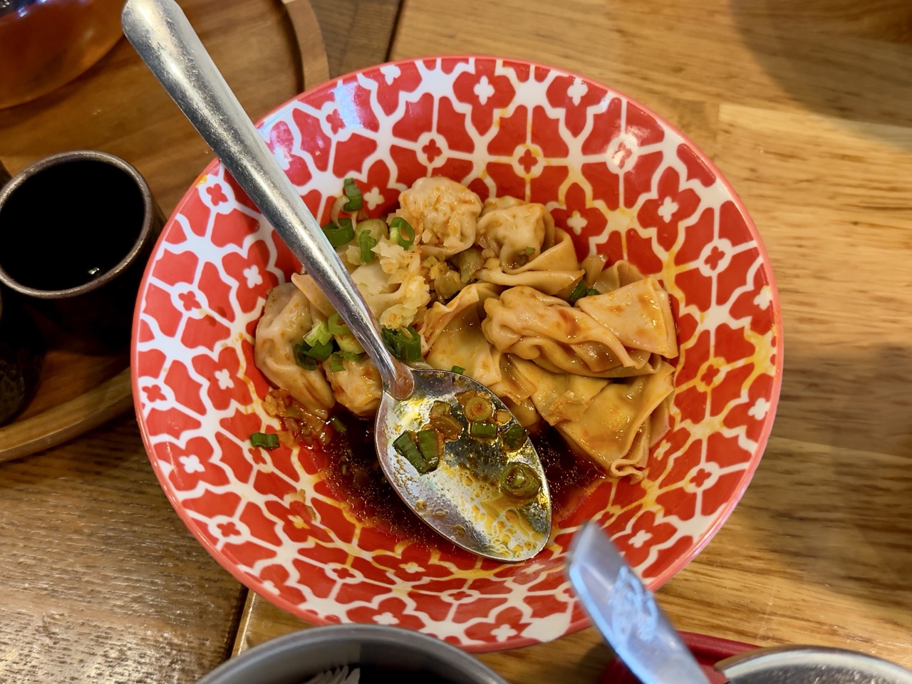 Overhead view of a red-and-white patterned ceramic bowl containing folded wontons in red chili oil, garnished with chopped scallions and minced garlic, alongside a metal serving spoon.