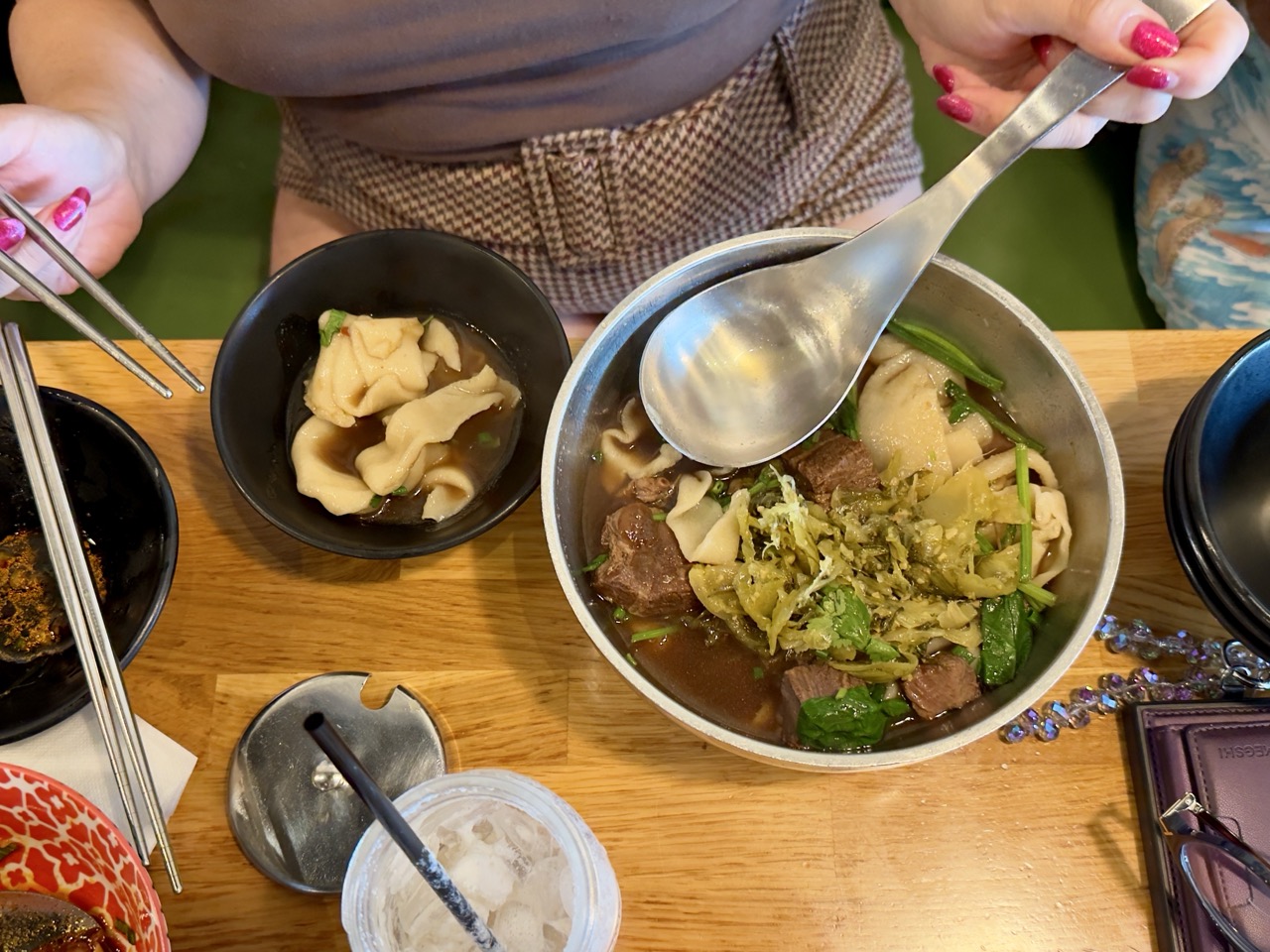 Overhead view of a person holding metal chopsticks and a large soup spoon over a metal bowl of Taiwanese beef noodle soup topped with pickled mustard greens, with a smaller black bowl of wide ribbon noodles beside it.