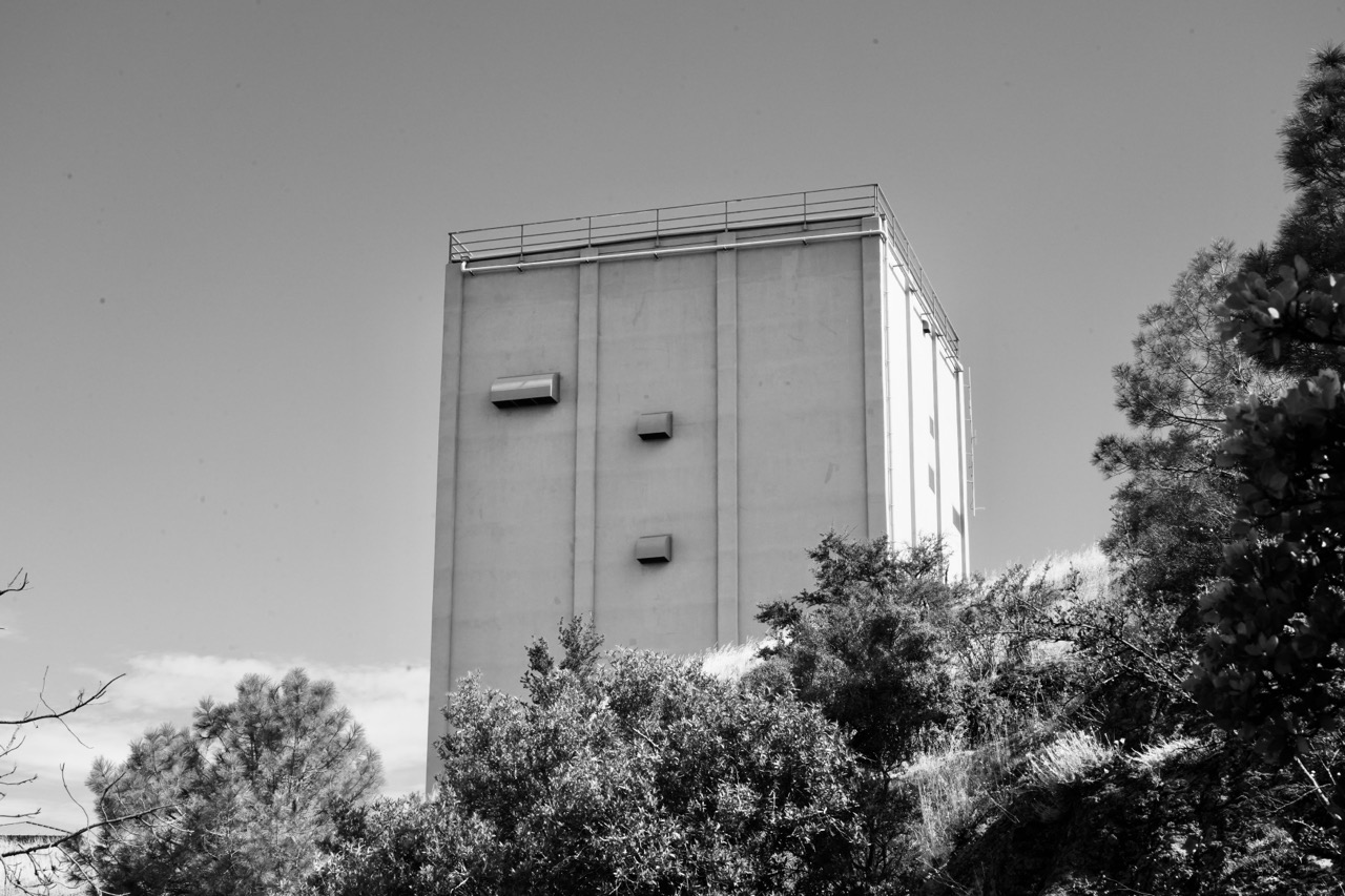 A black and white photograph of a tall, rectangular concrete tower surrounded by trees and brush on a hillside.