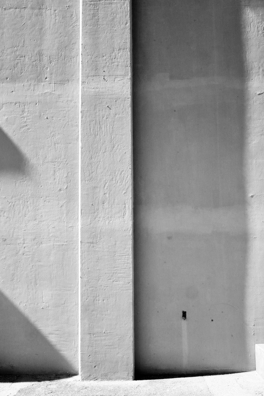 A close-up black and white view of a textured concrete wall sectioned by a vertical pillar and casting long geometric shadows.