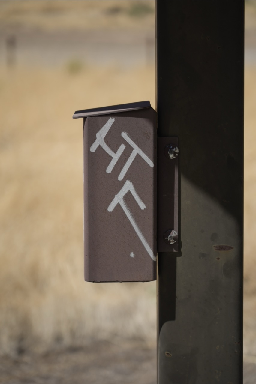 A vertical, close-up shot of a small, dark metal enclosure mounted to a structural post against a blurred, arid background, featuring white painted geometric markings on its side.