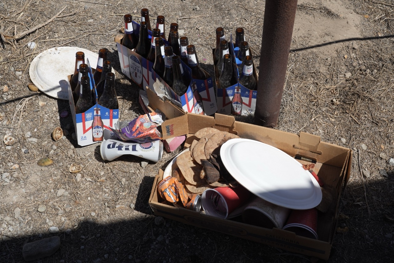 A high-angle view capturing discarded items on dry ground, including multiple cardboard cartons of bottled beer, white paper plates, red paper cups, a crushed can, and dried vegetation.