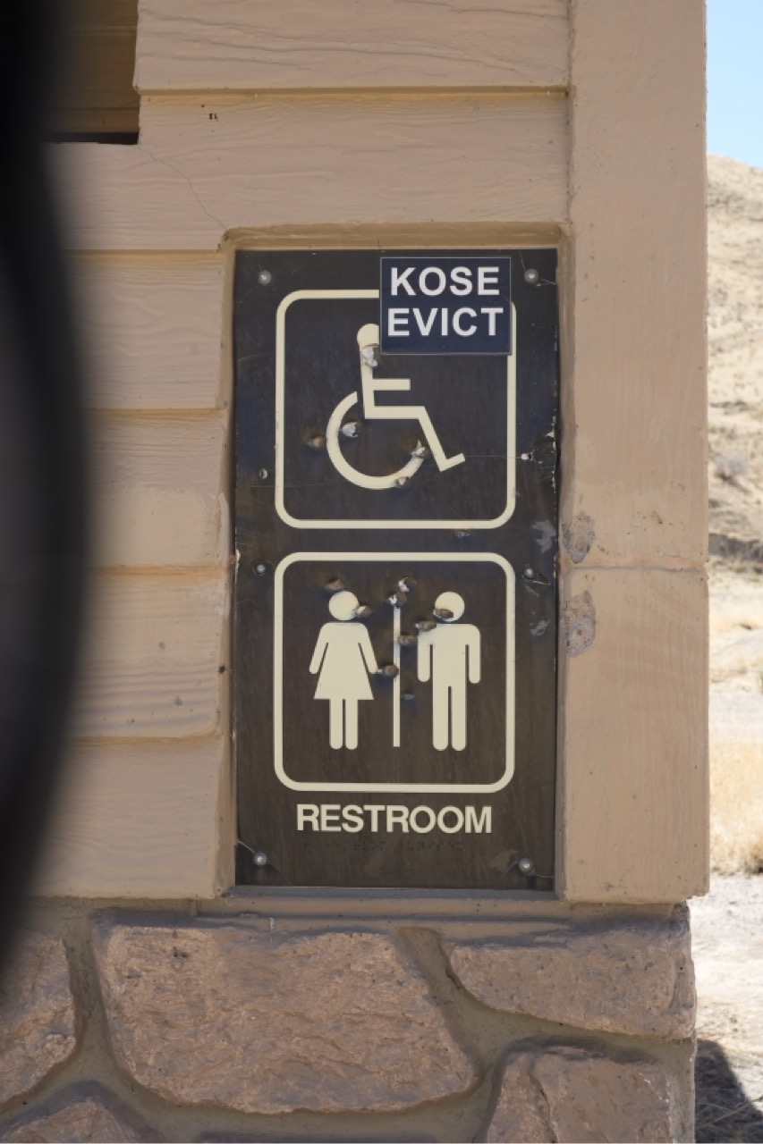 A close-up, vertical photograph of a restroom sign mounted on a textured wall, showing standard accessibility and gender-neutral or men’s/women’s restroom icons, with small sticker labels (KOSE EVICT) and bullet impacts or surface damage visible across the plate.